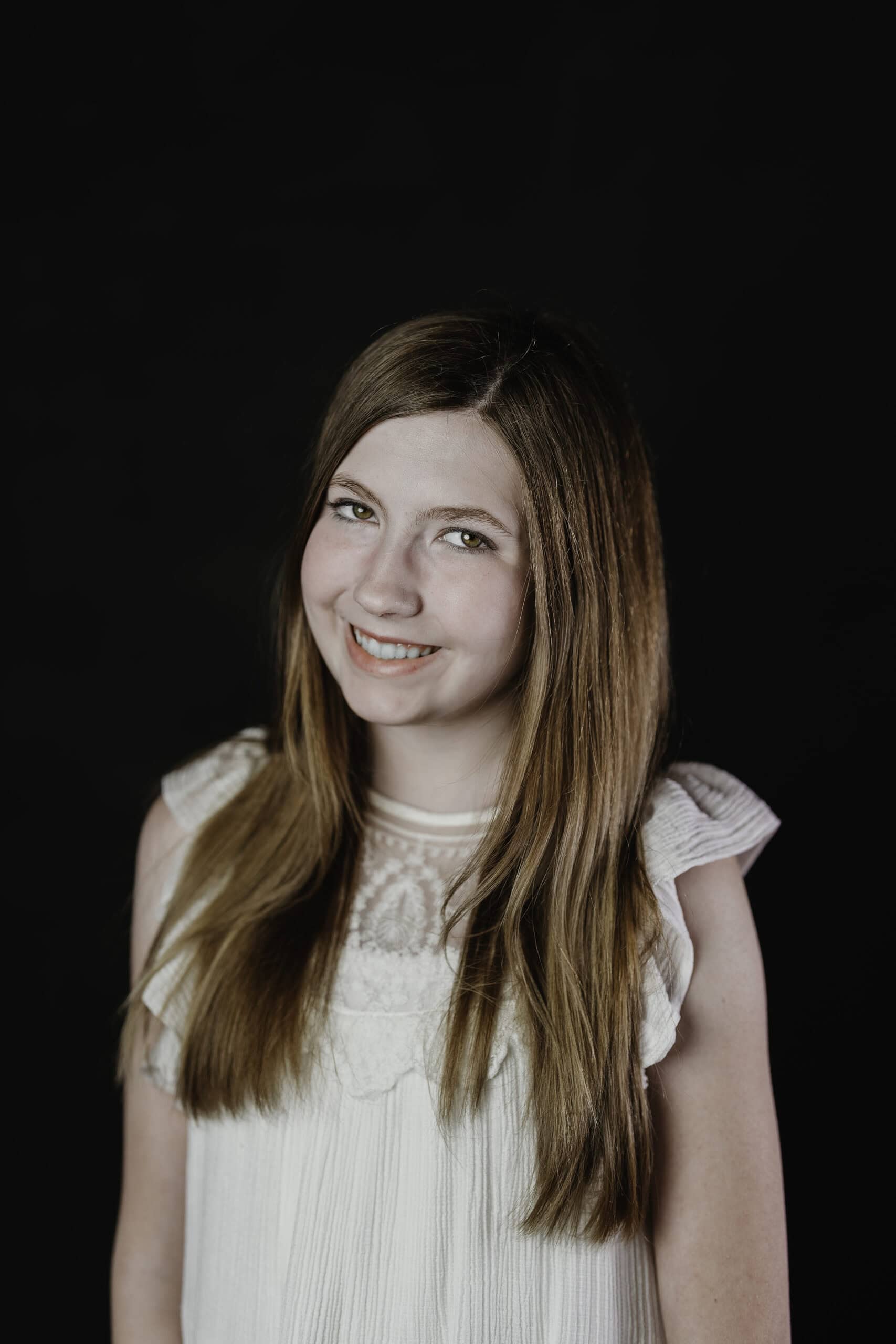Children's theater headshot of a smiling young actress with long hair in a white top on a black backdrop, by Captured in VA.