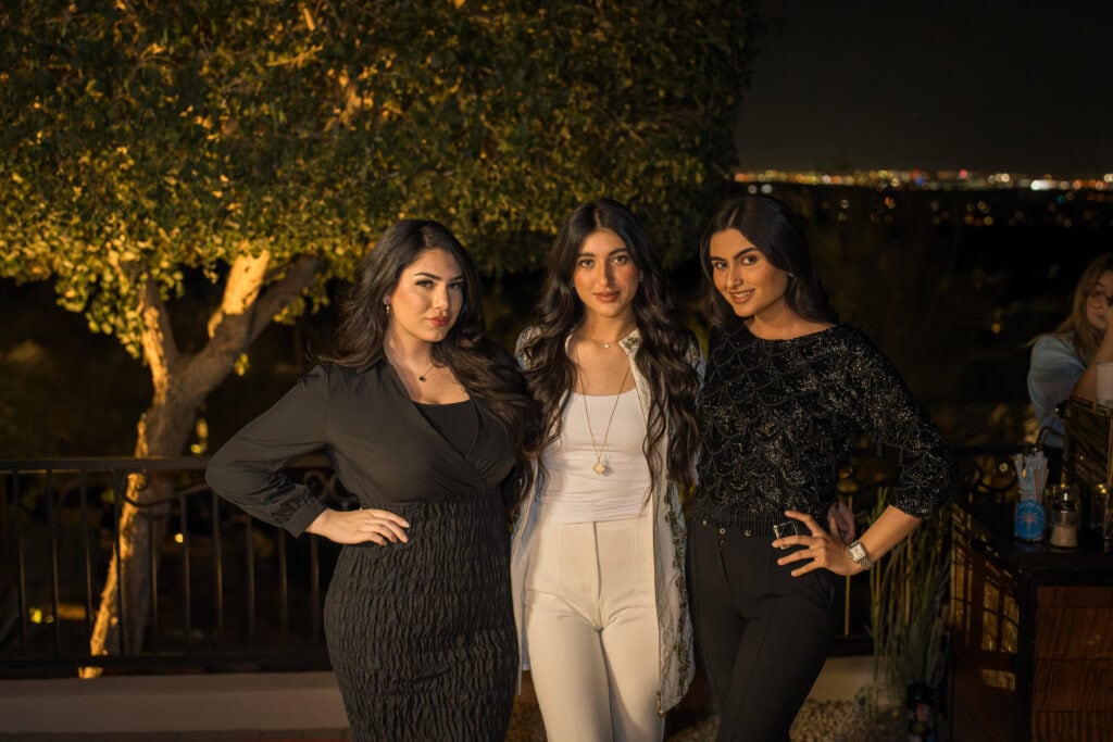 Three well-dressed women posing together on a terrace with city lights behind them at a nighttime birthday party, event photography by Captured in VA.