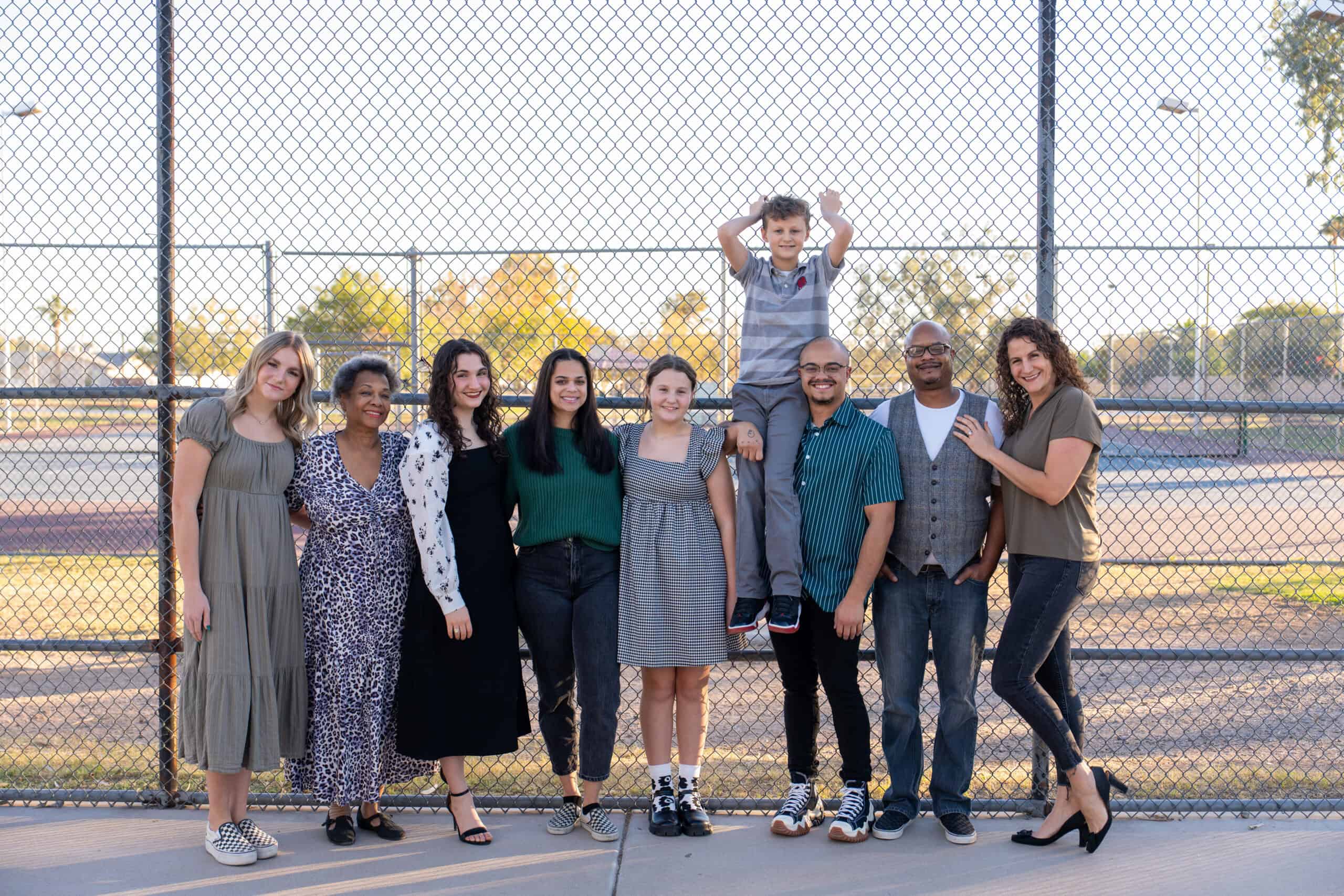 Extended family of nine smiling together along a chain-link fence at a park at golden hour, a young boy perched up top, by Captured in VA.