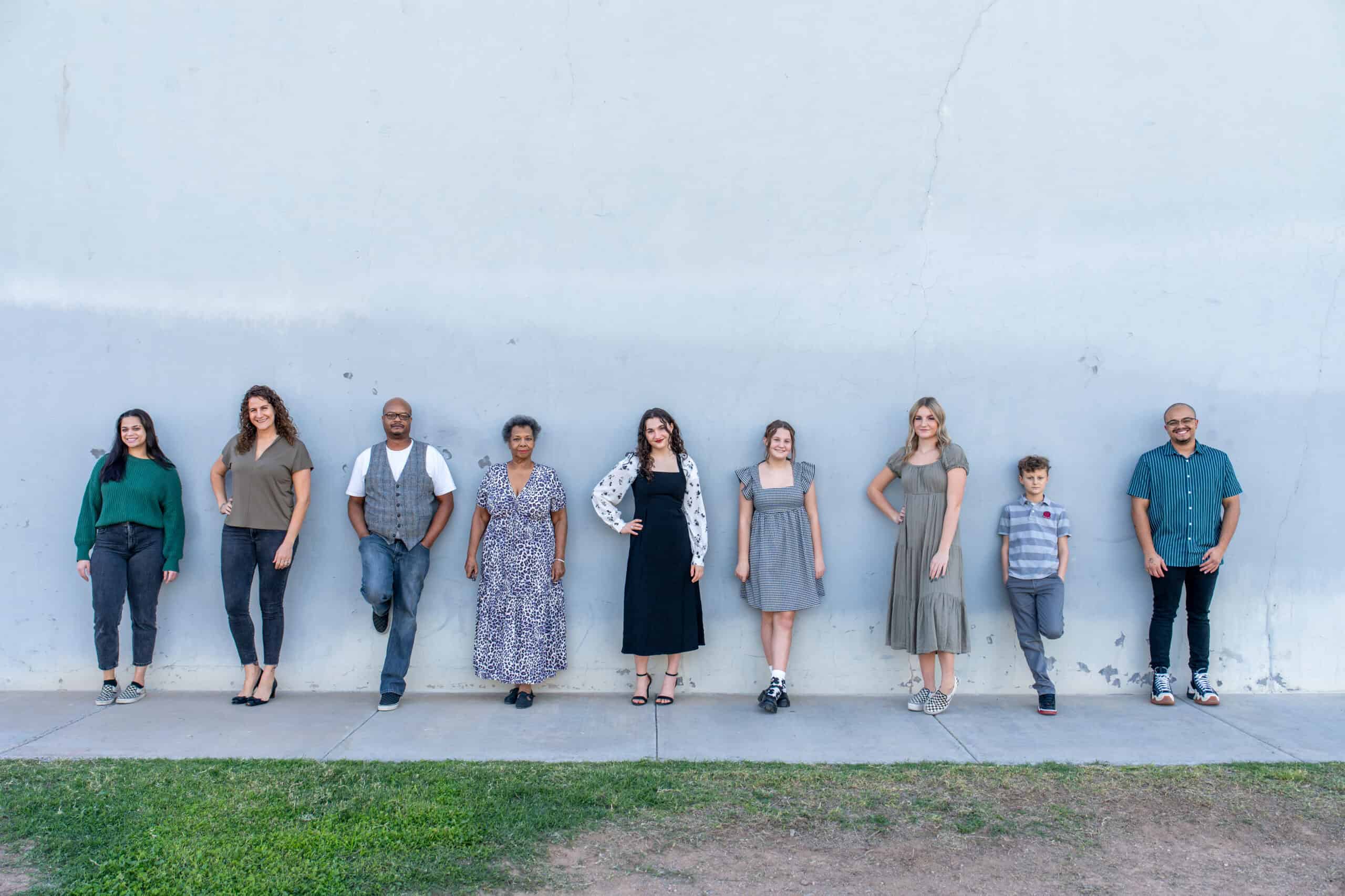 Extended family of nine lined up and leaning against a light stucco wall in a relaxed row, a family portrait by Captured in VA.