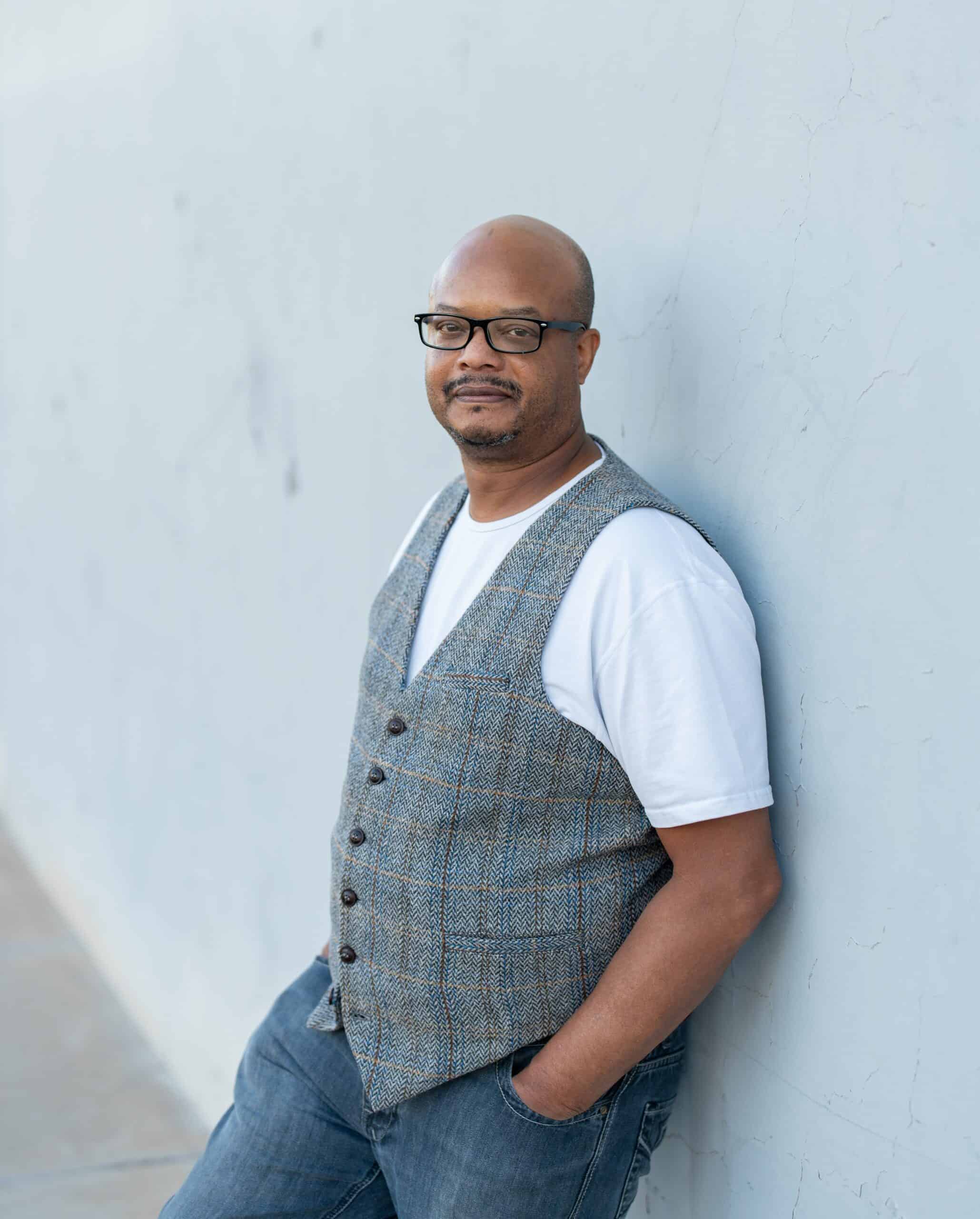 Relaxed individual portrait of a man in a tweed vest and white tee leaning against a wall, from a family session by Captured in VA.