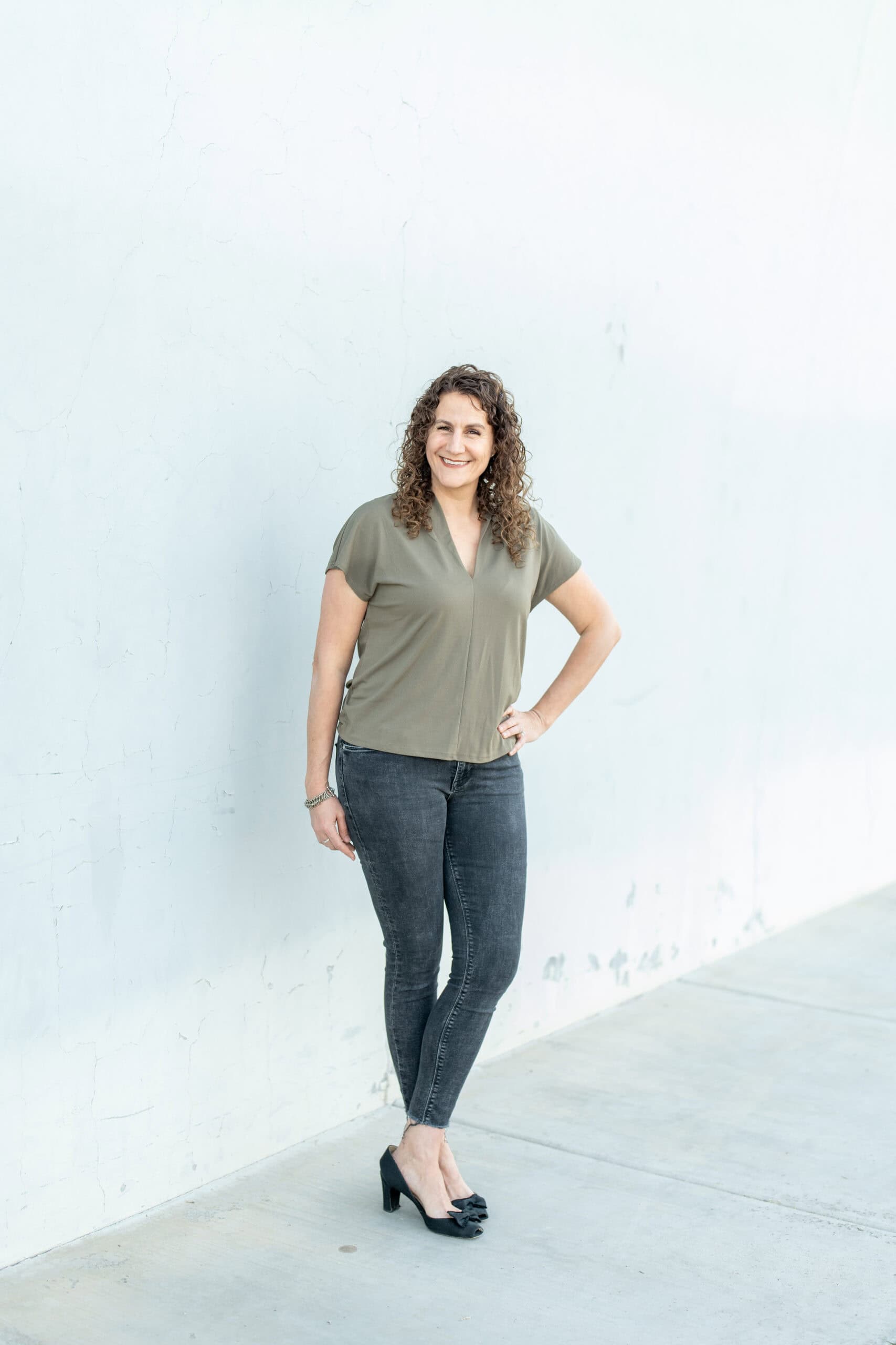 Full-length individual portrait of a smiling woman with curly hair in an olive top and dark jeans against a white wall, by Captured in VA.