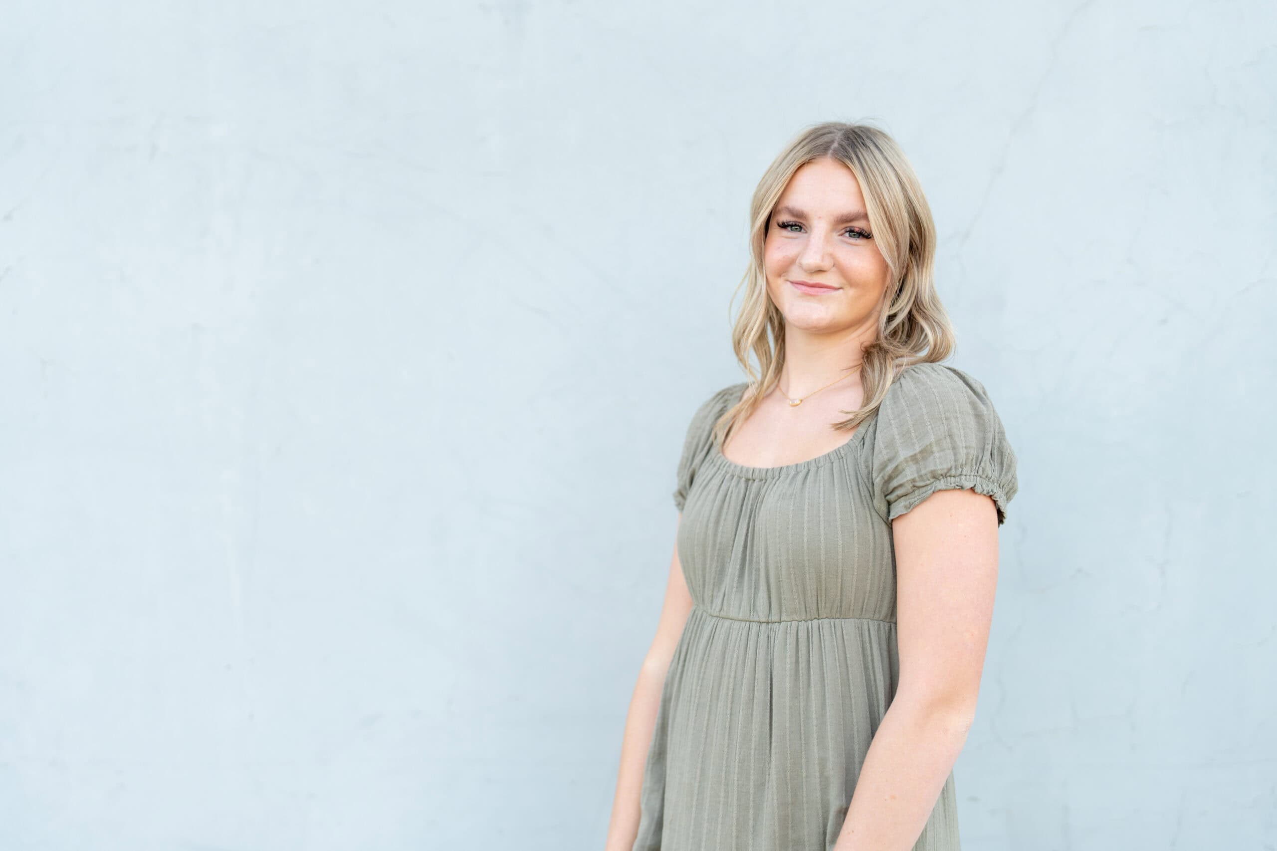 Individual portrait of a young woman with blonde hair in a sage puff-sleeve dress smiling against a light wall, by Captured in VA.