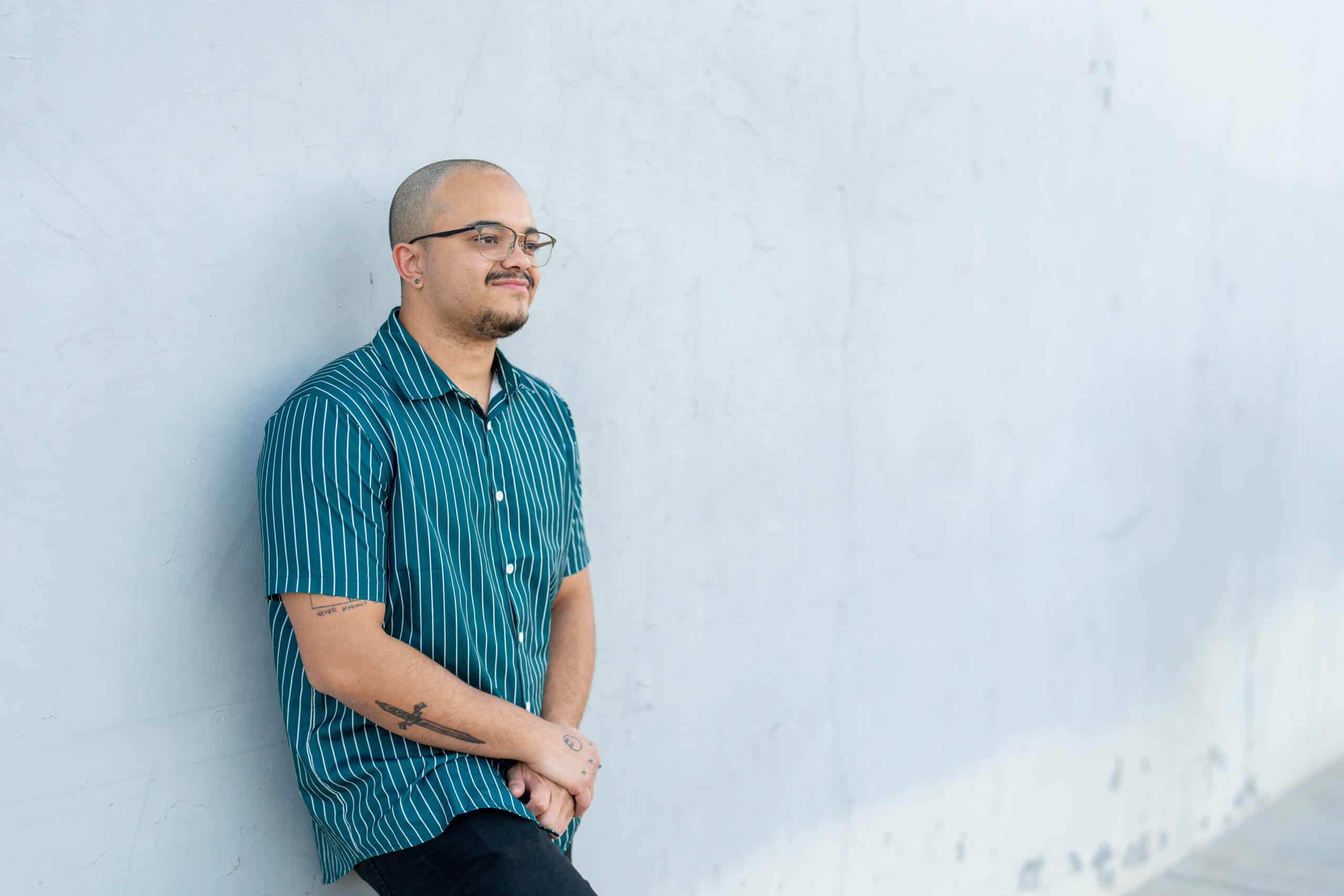 Individual portrait of a man in a green striped shirt and glasses leaning against a white wall, looking off-camera, by Captured in VA.