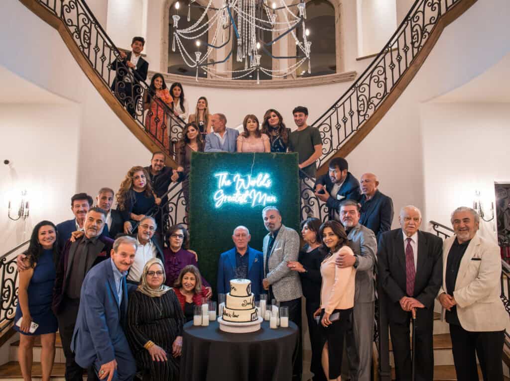 Large family gathered on a grand staircase around a tiered cake and 'World's Greatest Man' sign for a milestone birthday celebration, event photography by Captured in VA.