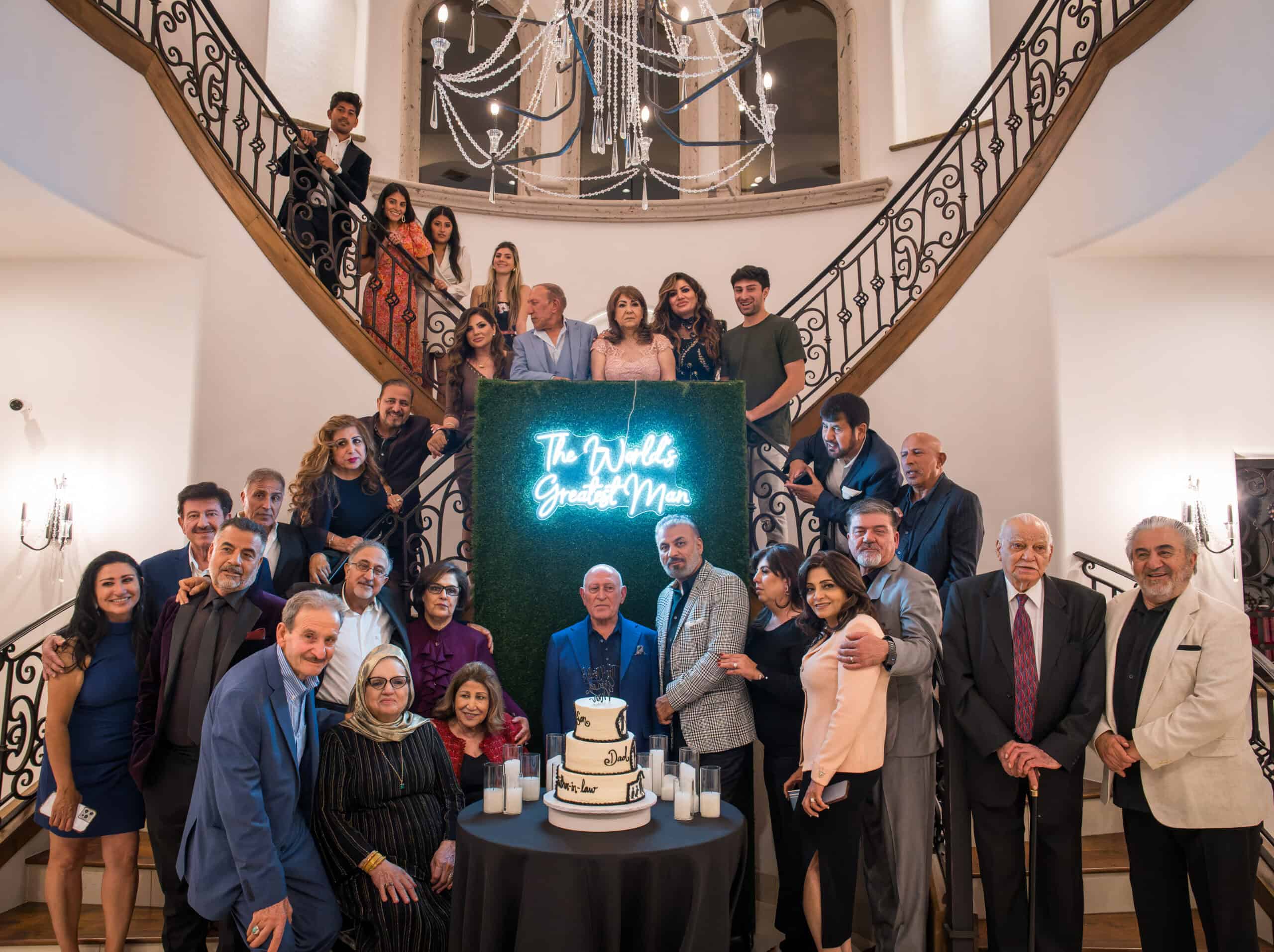 Large family gathered on a grand staircase around a tiered cake and 'World's Greatest Man' sign for a milestone birthday celebration, event photography by Captured in VA.