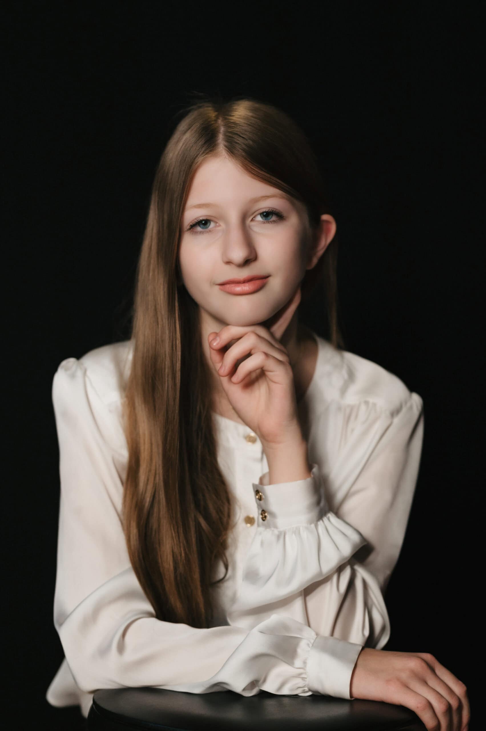 Studio portrait of a young girl with long hair in a white ruffled blouse, hand to chin against a black backdrop, by Captured in VA.