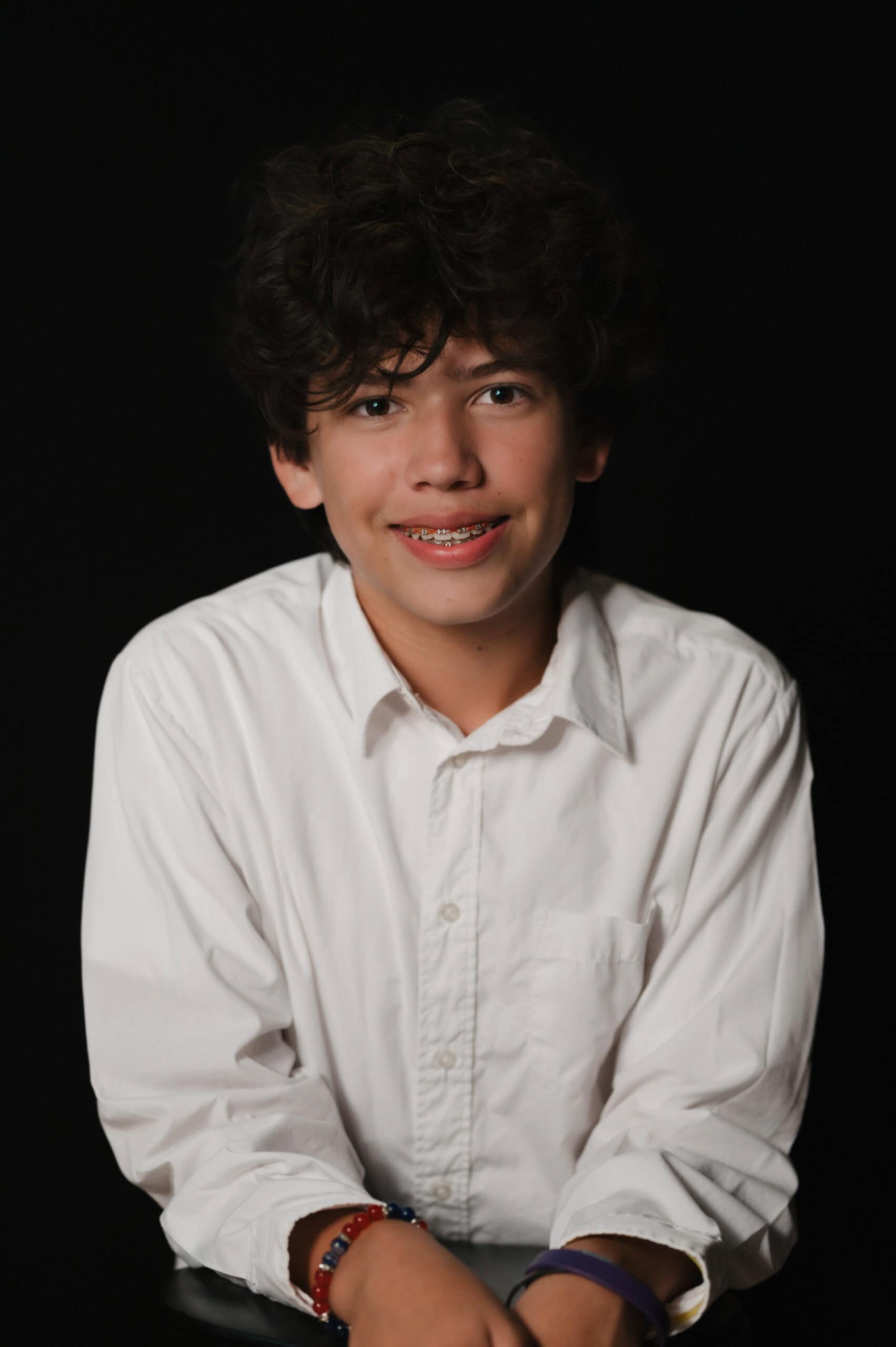 Studio portrait of a smiling teen boy with curly hair in a white button-down shirt against a black backdrop, by Captured in VA.