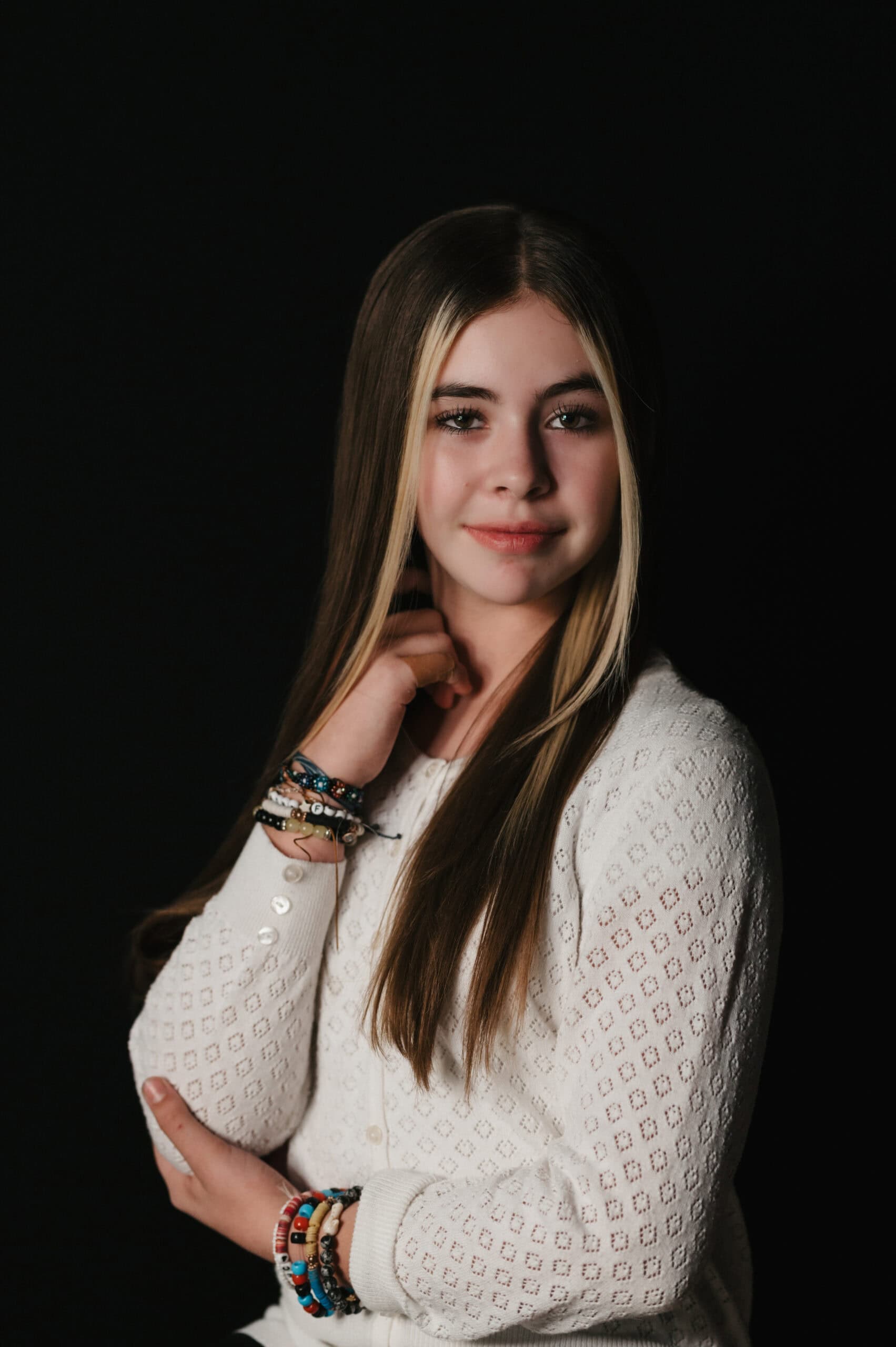 Studio portrait of a young girl in a white pointelle-knit sweater with stacked bracelets against a black backdrop, by Captured in VA.
